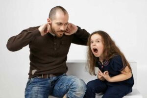 Angry little girl with long loose hair screams, misbehaves because her father won't let her watch cartoons. Frustrated young bearded man plugs his ears, can't stand his daughter's annoying screams / Freepik