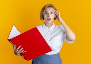 young surprised russian blonde puts her hand on her head holding a file folder isolated on an orange background with a copy space / Freepik