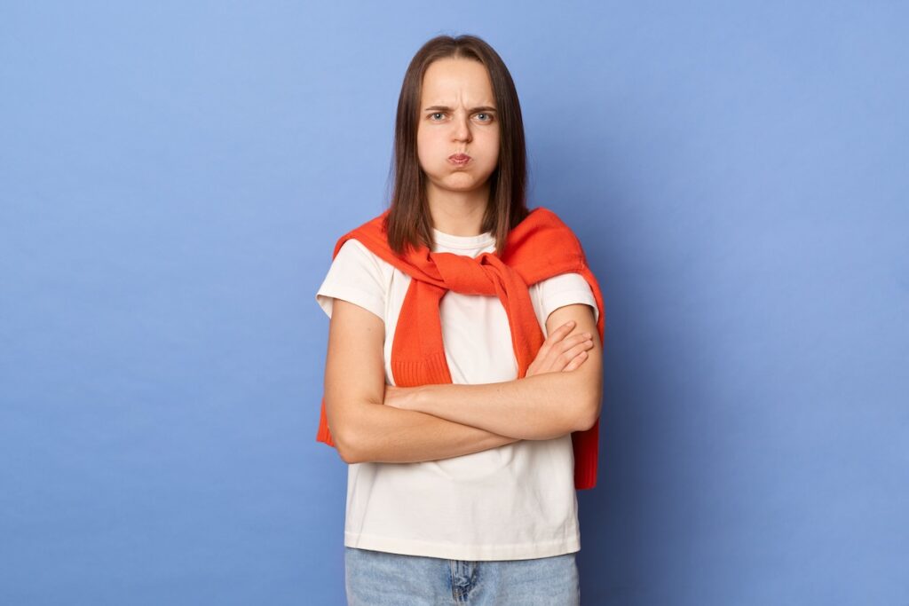 Cнимок грустной расстроенной девушки, которая дует в щеки, обиженная на кого-то; женщина в белой футболке и оранжевом свитере, повязанном на плечах, стоит у синей стены