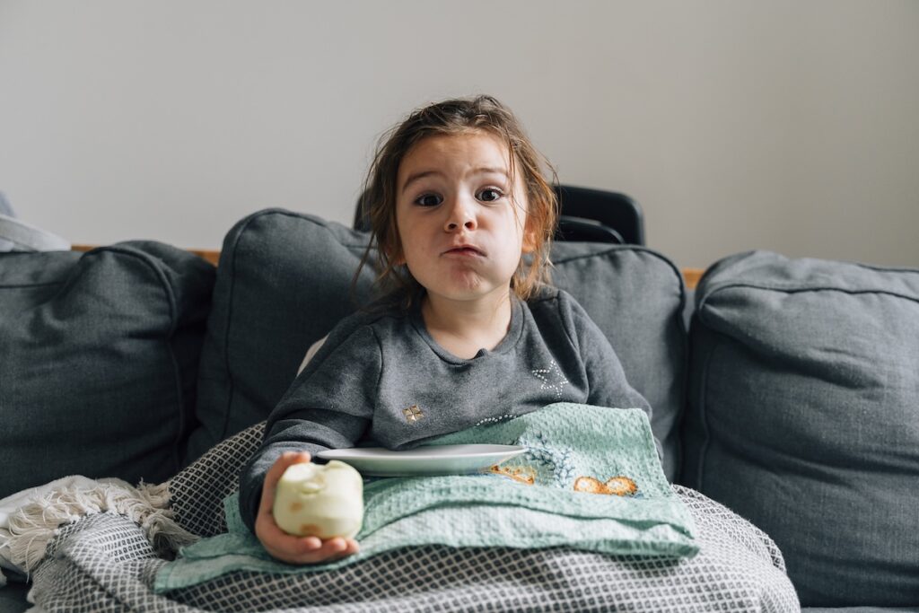 Девочка дошкольного возраста ест яблоко на диване в гостиной.