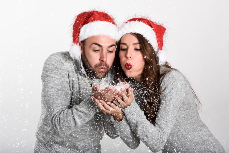 Young happy couple in winter clothes and Santa hat blowing snowflakes off their hands on white background.