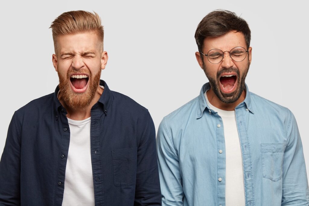 Photo of irritated guys, shouting loudly, with wide open mouths, trendy hairstyles and stubble, standing shoulder to shoulder on a white background. People, irritation and negative facial expressions.