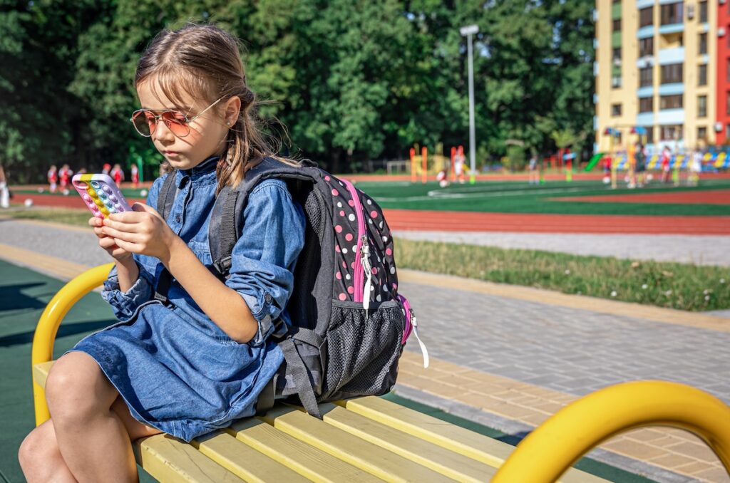 An elementary school student with a backpack in hand uses a smartphone while sitting near the school.