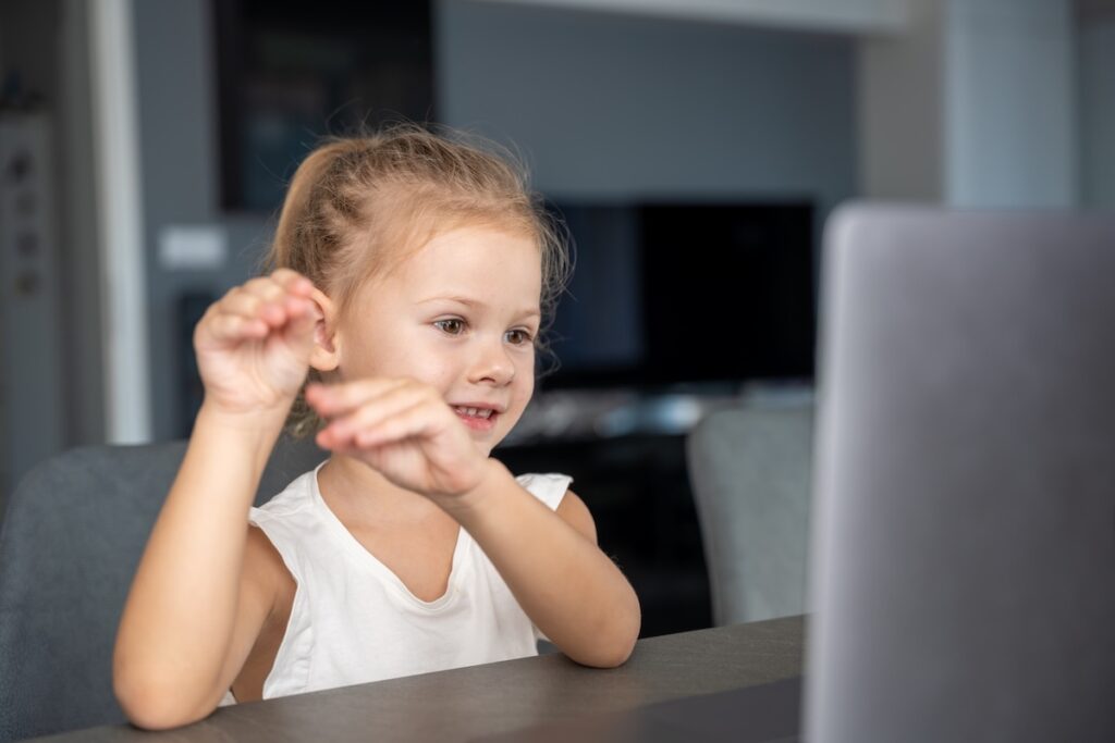 Маленькая девочка занимается пальчиковой гимнастикой онлайн на ноутбуке дома. Концепция дистанционного обучения.