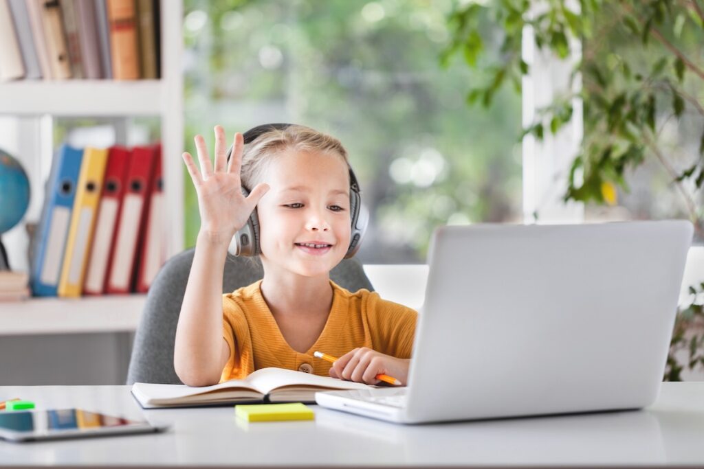 Улыбающийся маленький ребенок в наушниках участвует в видеозвонке на дистанционном занятии, используя ноутбук.