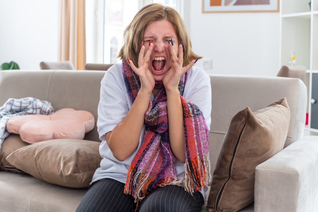 A young woman with a sickly look, wearing a warm scarf around her neck, feeling terrible about the virus, screams in panic as she sits on a couch in a bright living room.