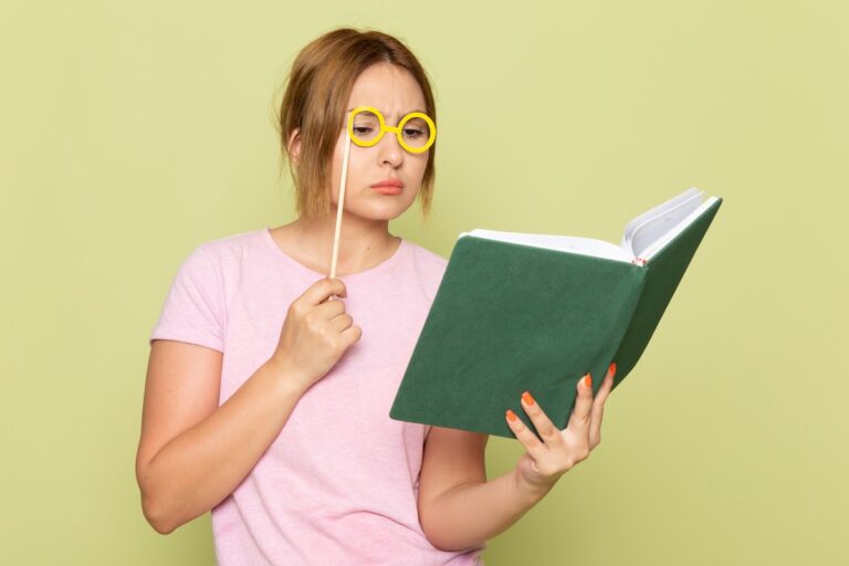 Front view: young beautiful girl in pink t-shirt and blue jeans posing with toy sunglasses, reading a book on green background. Woman, lady, beauty.