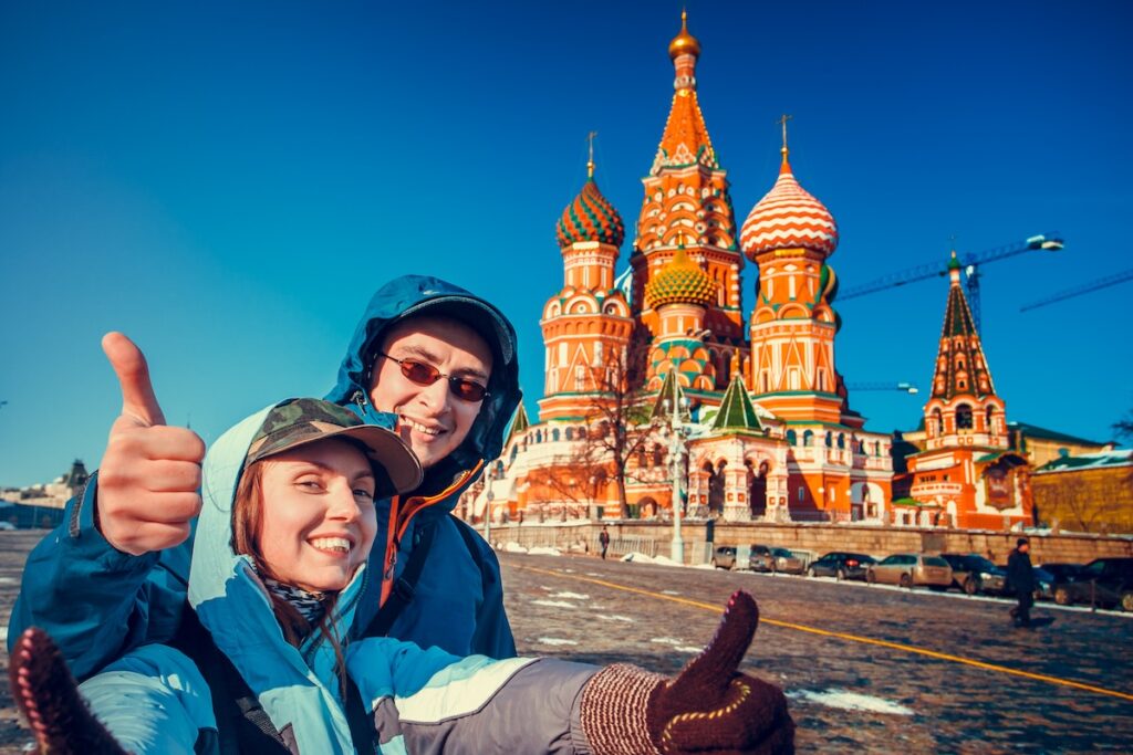 Счастливые туристы осматривают город, поднимая пальцы вверх, рядом с собором Василия Блаженного. Красная площадь, Москва, Россия. Праздник, путешествие, отдых. Зимний сезон, яркие краски, чистое голубое небо.