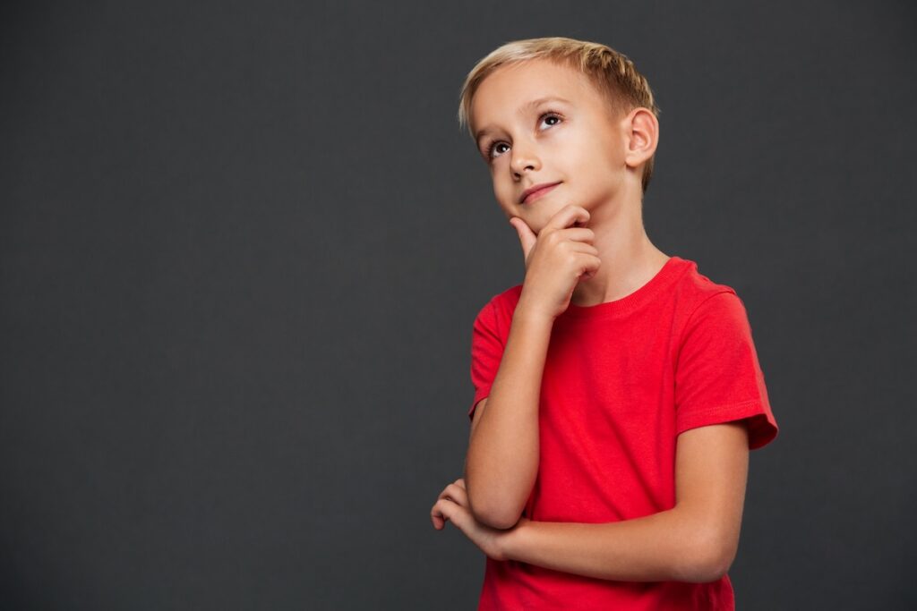 Image of a pensive, sweet little boy standing on a gray background. He is looking away.