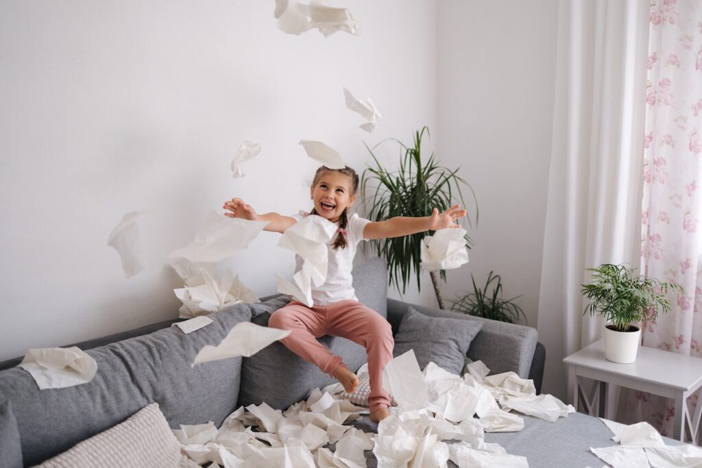 Очаровательная маленькая девочка устраивает беспорядок дома. Пятилетняя девочка разбрасывает салфетки по гостиной. Фотография высокого качества.