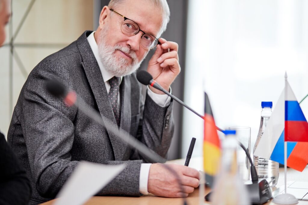 Пожилой руководитель, политик в костюме сидит на конференции, слушает мнения людей и обменивается идеями, с микрофоном в офисе. Международная встреча, саммит.