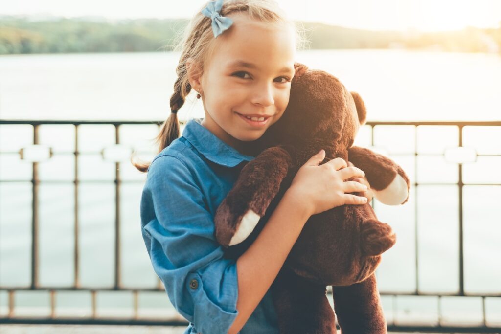 Крупный план девочки, обнимающей милого плюшевого медведя и смотрящей в камеру на фоне озера и деревьев. На фото — девочка в джинсовом платье. Концепция детской дружбы. Счастливый улыбающийся ребенок.