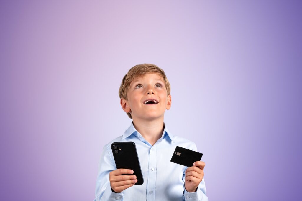 Улыбающийся школьник с телефоном и кредитной картой в руках, смотрящий вверх. Мечтательный портрет на пустом фиолетовом фоне. Концепция мобильного приложения и онлайн-шопинга.