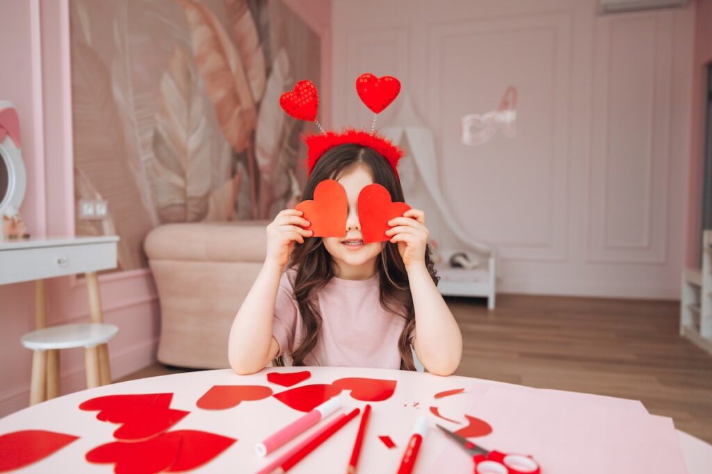 Маленькая девочка держит красные карточки в форме сердца и закрывает ими глаза. День святого Валентина.