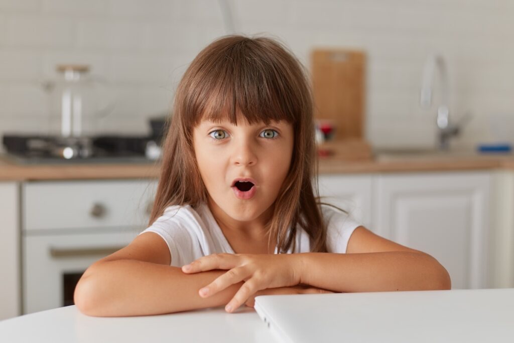 A little girl sits at home at her desk with her laptop closed. The cute little girl looks at the camera with her mouth open expressing shock, the dark-haired girl is surprised to see something unexpected.