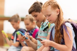 Elementary education, friendship, childhood, technology and people - a group of happy elementary school students with smartphones and backpacks sit on a bench outside.