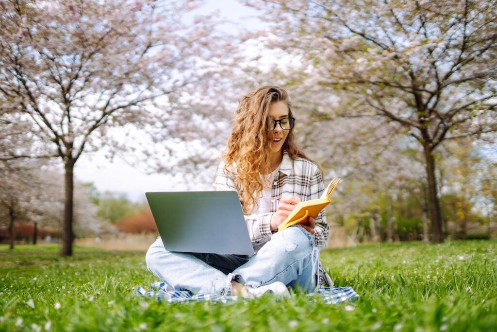 Женщина сидит на зеленой траве возле цветущих деревьев, с ноутбуком в руках, что-то записывает в блокнот. Концепция планирования.