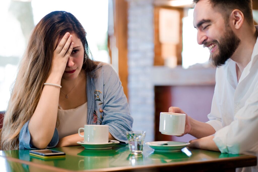 A girl holds her head in her hand, a man smiles, they are sitting at a table with cups.
