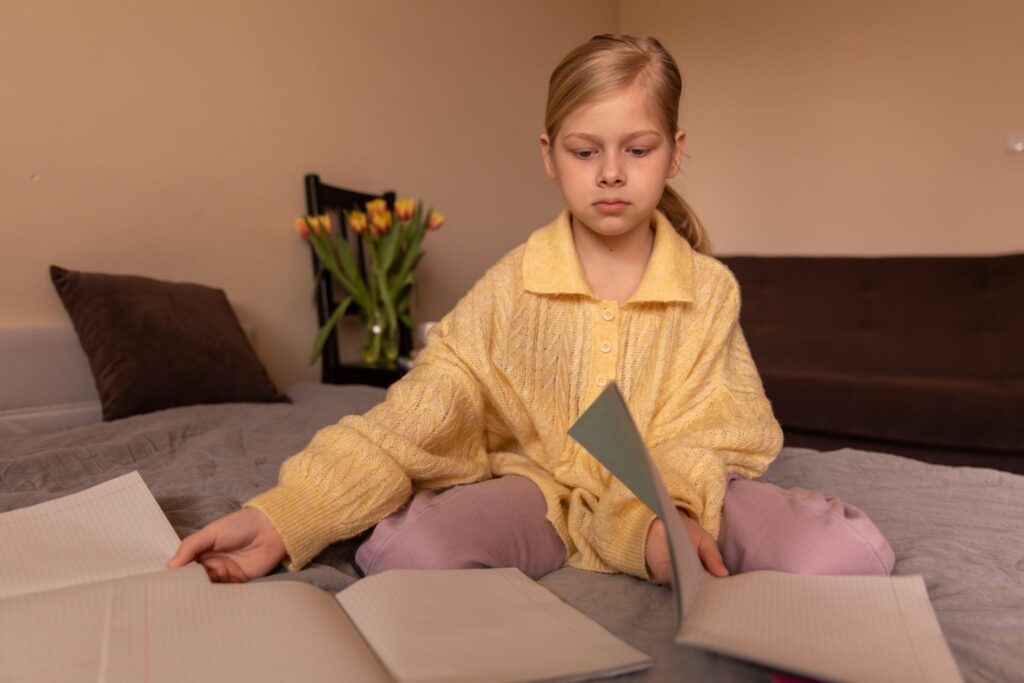 A girl sits on the bed with open notebooks.