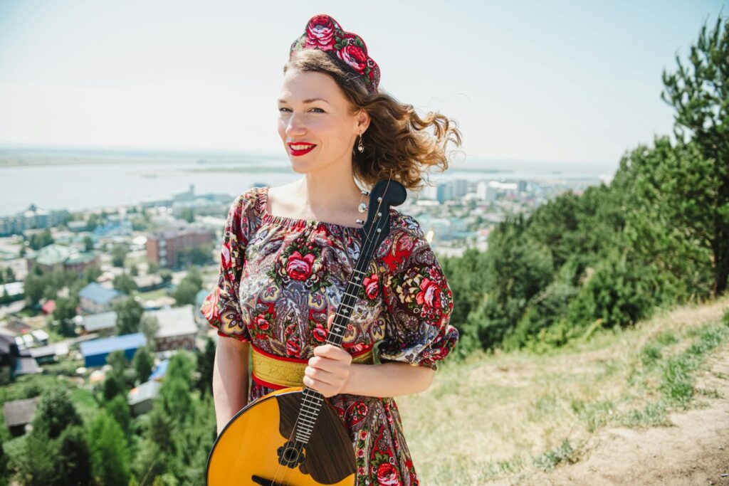 A girl on a mountain with a city in the background, wearing a kokoshnik with flowers, holding a domra.