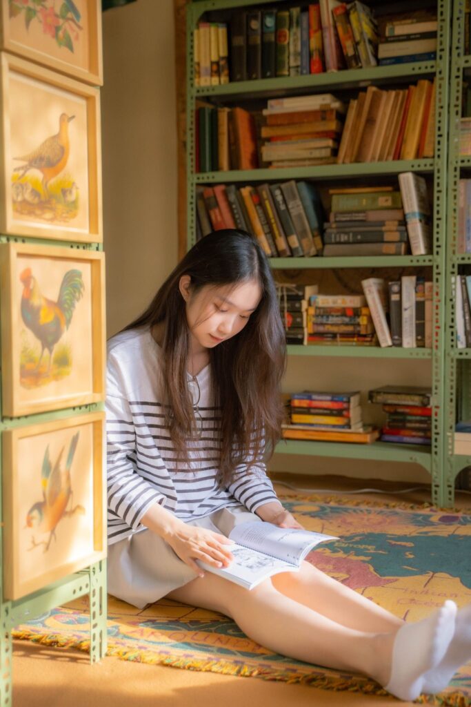 Девочка-подросток сидит на полу с книгой в комнате с книжными полками, образ тихого чтения в возрасте первых влюблённостей