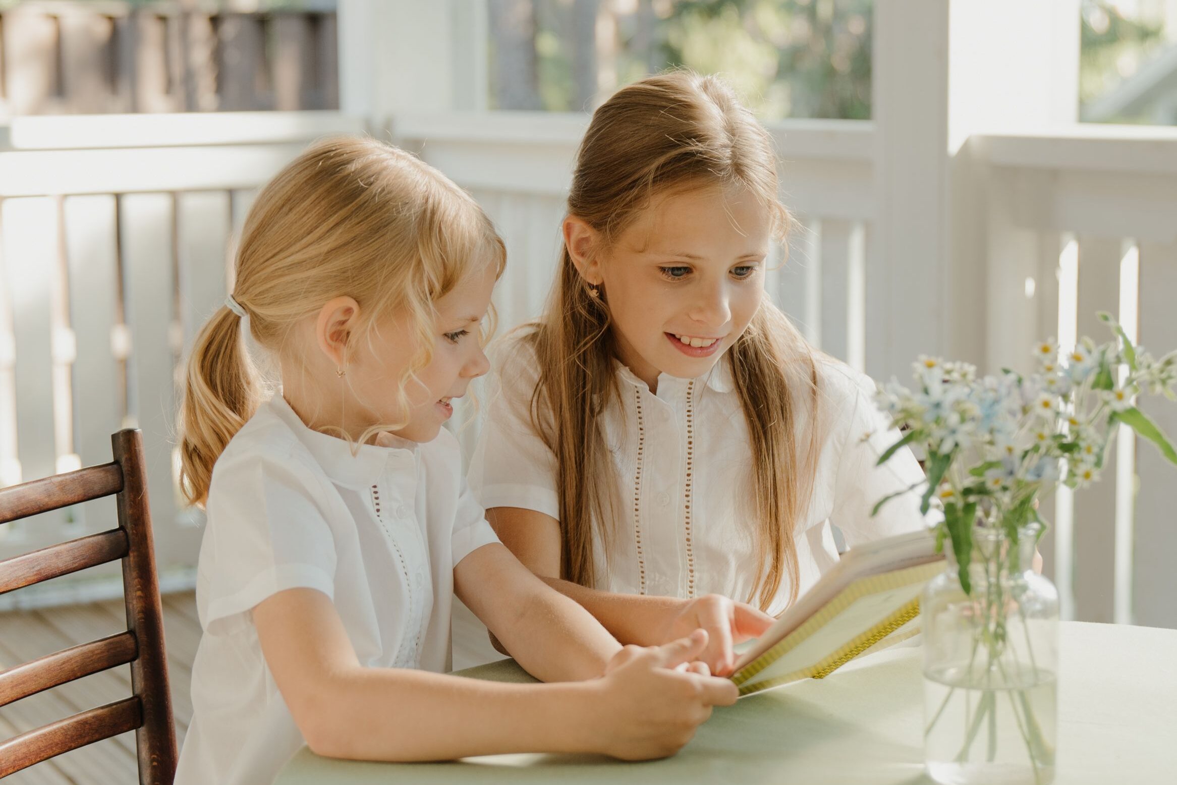 Two young girls reading a book together on a sunlit porch, surrounded by nature.