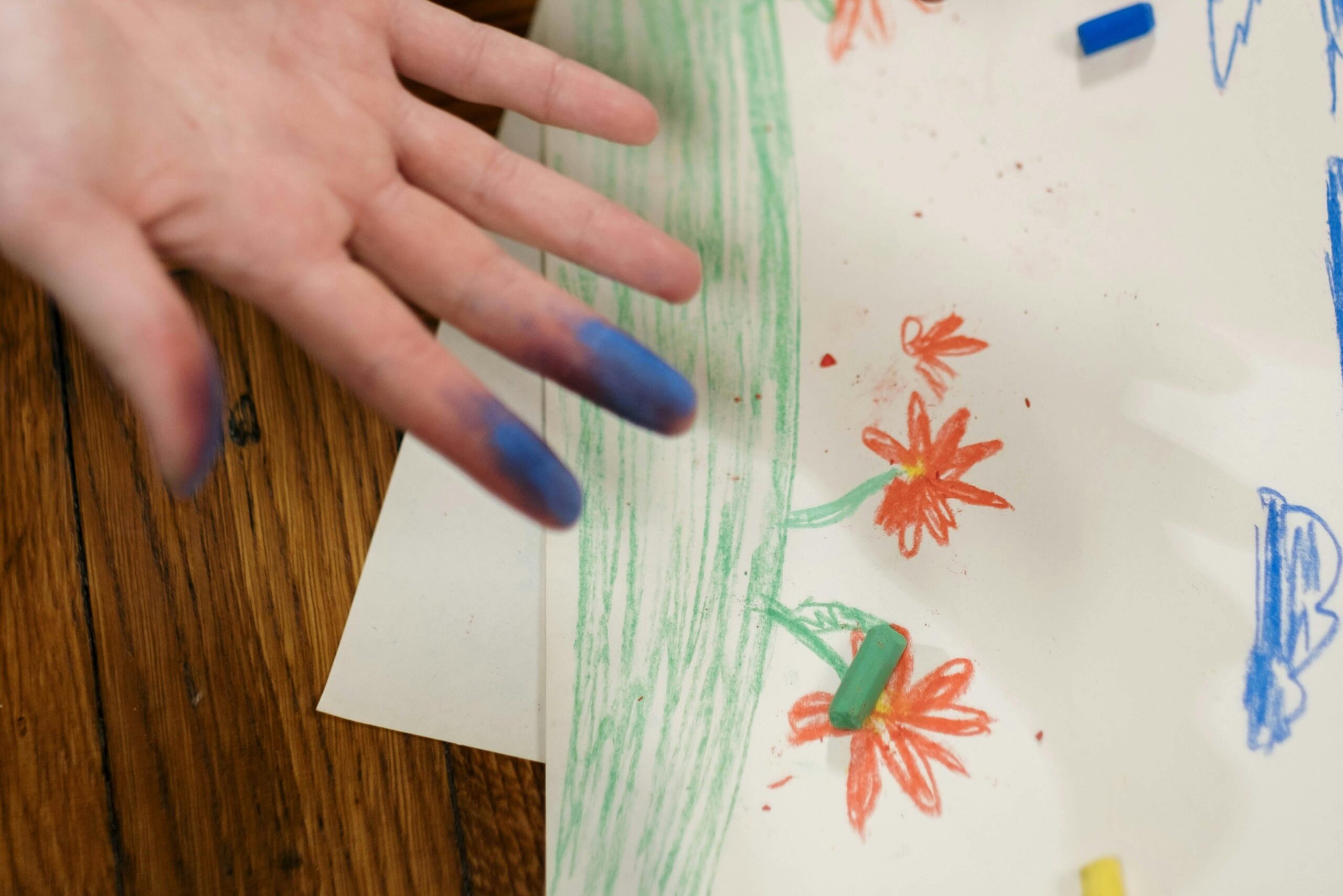 A child's hand with crayons and child's drawing in the background