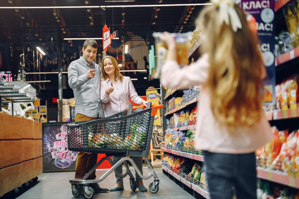 Family in a grocery aisle, young daughter holding a snack box and asking her parents for it, wide framing, child not close-up