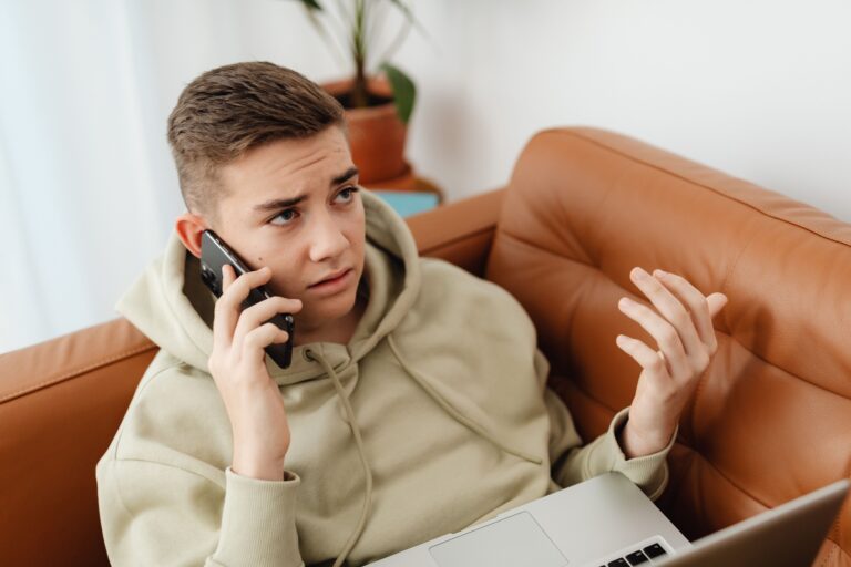 A young man is talking on the phone while working on a laptop; he looks concerned and frustrated.