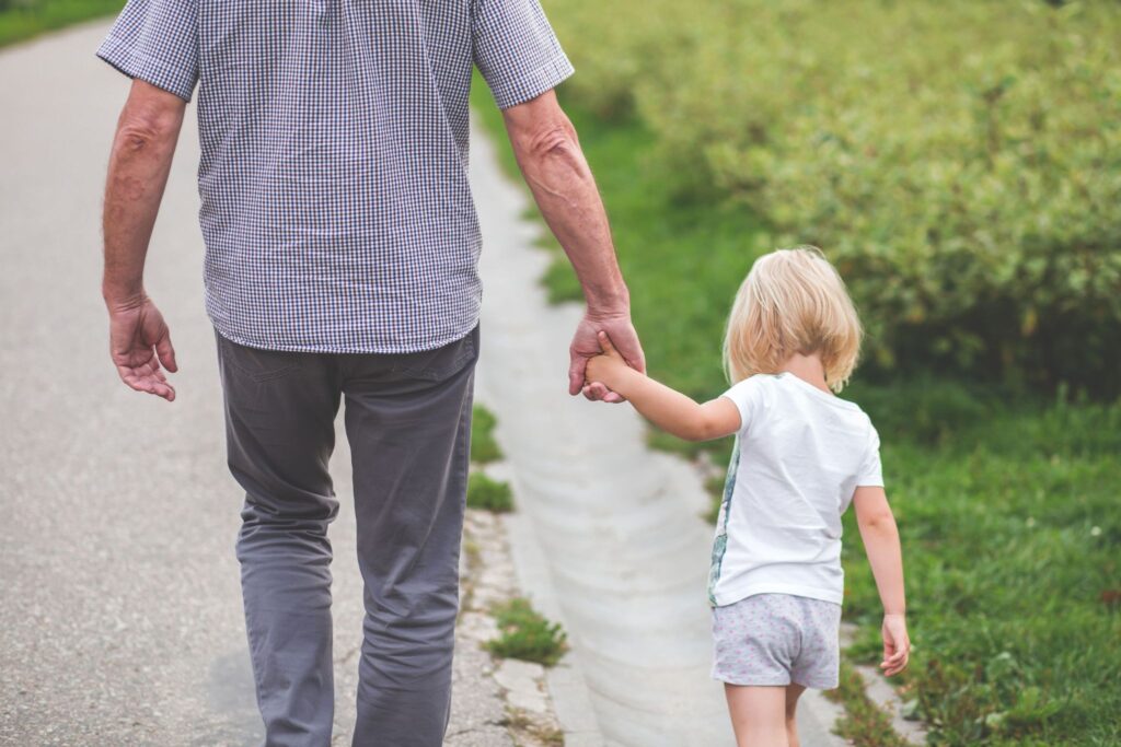 An adult and a child walking hand-in-hand in a park, seen from behind